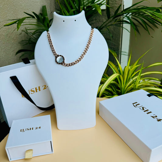 Elegant gold chain necklace with a chunky curb-link design, featuring a single circular pendant with a faceted crystal accent. The minimalist yet bold style makes it a versatile statement piece. Displayed on a white mannequin with luxury packaging in the background.