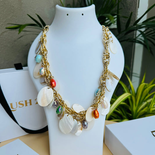A chunky statement necklace featuring gold-tone chain links adorned with white shell discs and colorful faceted beads. The vibrant mix of orange, blue, and iridescent accents creates a beach-inspired, elegant look. Displayed on a white jewelry bust with a lush green backdrop.