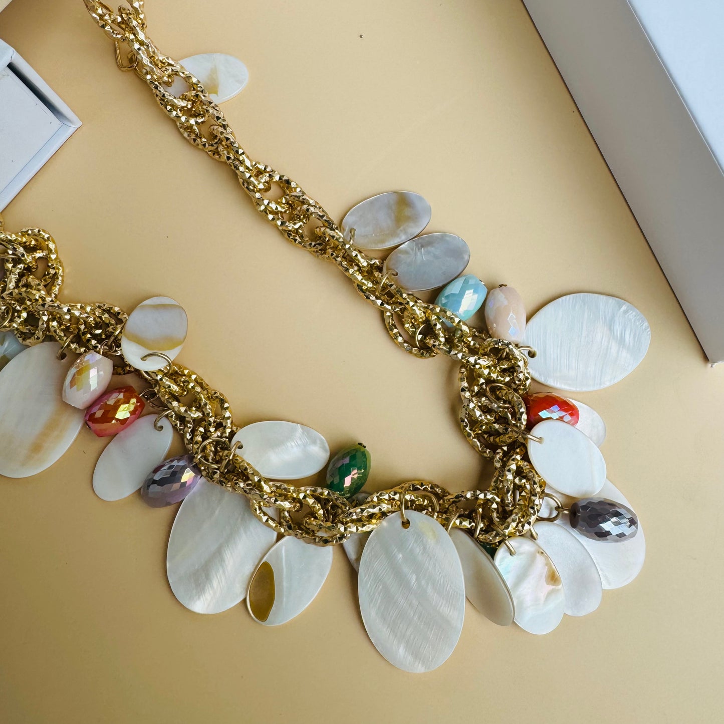 A chunky statement necklace featuring gold-tone chain links adorned with white shell discs and colorful faceted beads. The vibrant mix of orange, blue, and iridescent accents creates a beach-inspired, elegant look. Displayed on a white jewelry bust with a lush green backdrop.