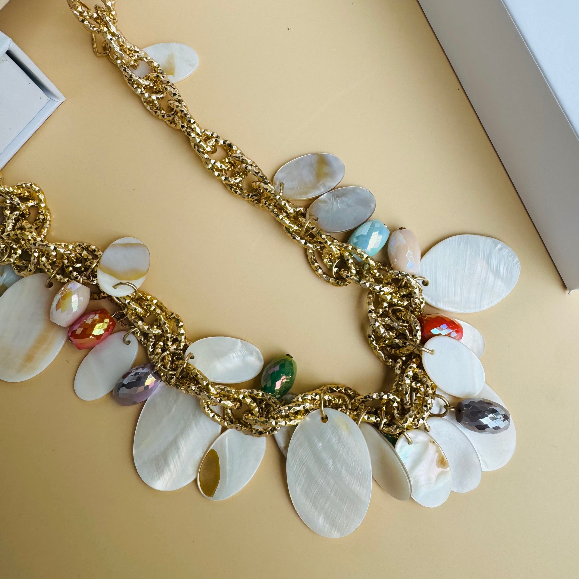 A chunky statement necklace featuring gold-tone chain links adorned with white shell discs and colorful faceted beads. The vibrant mix of orange, blue, and iridescent accents creates a beach-inspired, elegant look. Displayed on a white jewelry bust with a lush green backdrop.