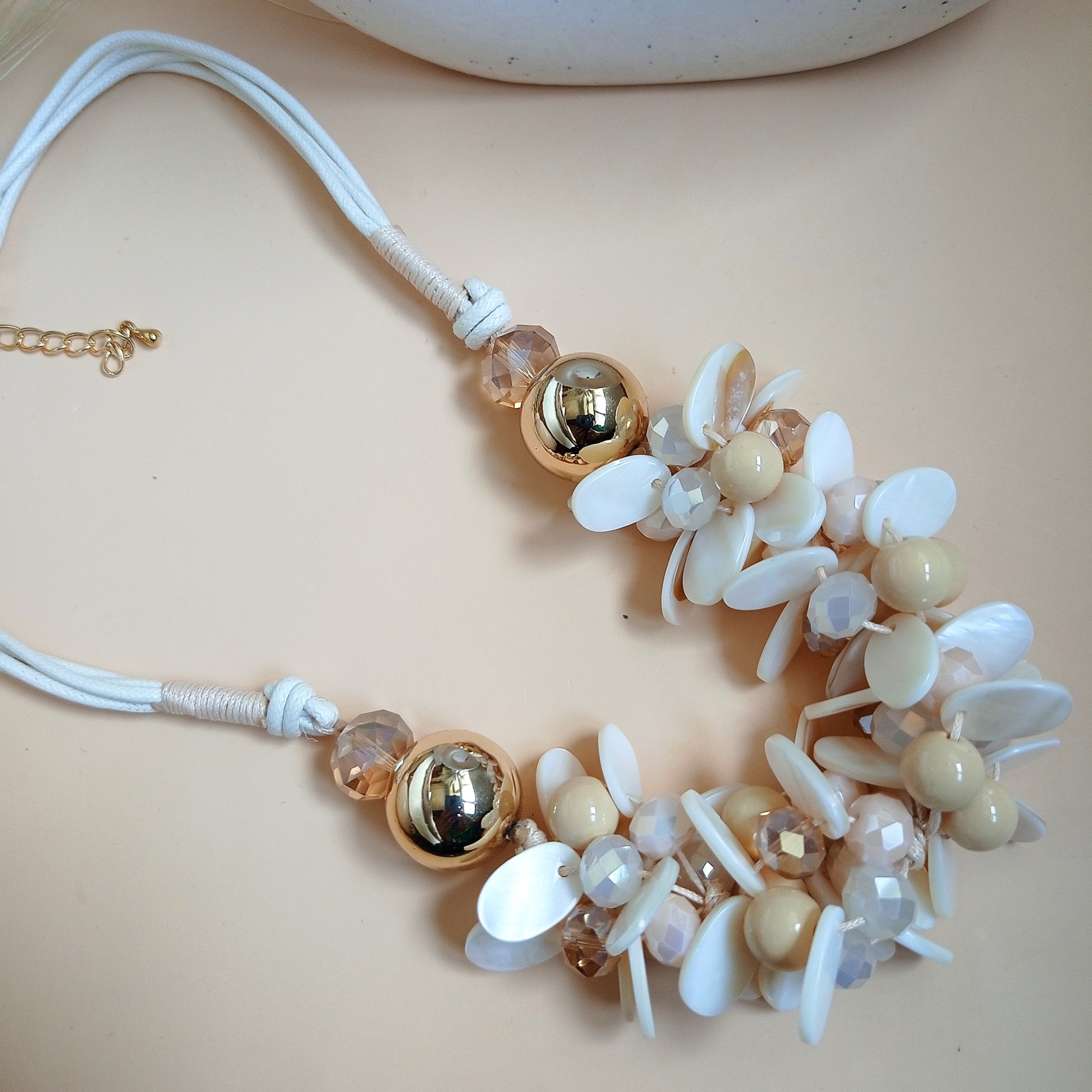 Statement necklace with white and beige floral shell beads, gold-tone accents, and a soft rope-style chain, displayed on a neutral background