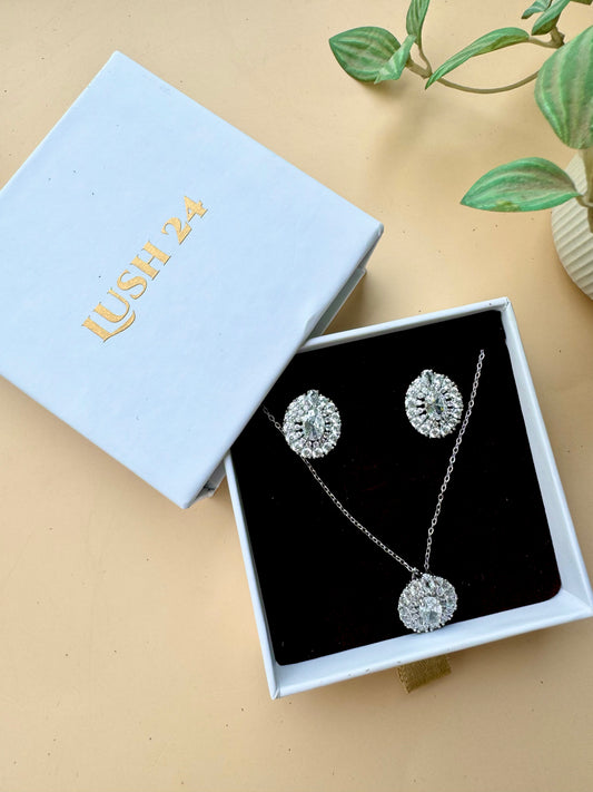 A set of sparkling silver-toned jewelry featuring flower-shaped designs with clear crystals. The set includes a pendant necklace and matching stud earrings, presented in a light blue LUSH 24 branded gift box with a black velvet insert.