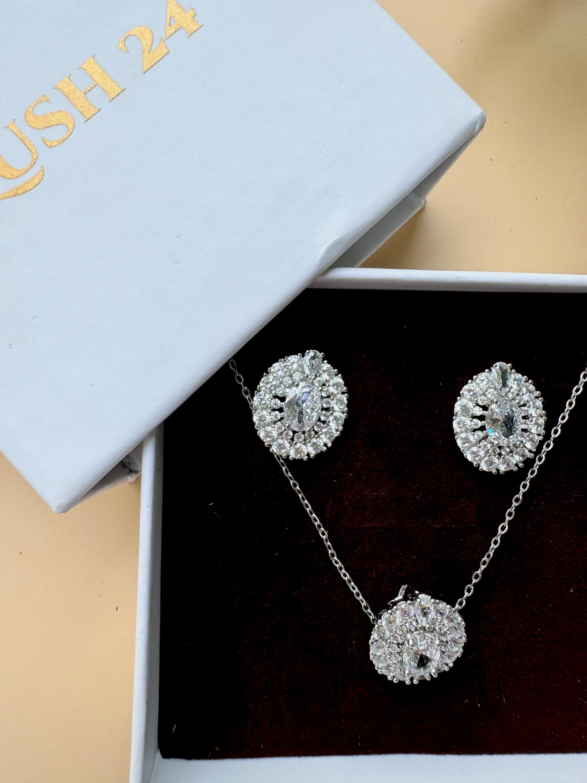 A set of sparkling silver-toned jewelry featuring flower-shaped designs with clear crystals. The set includes a pendant necklace and matching stud earrings, presented in a light blue LUSH 24 branded gift box with a black velvet insert.
