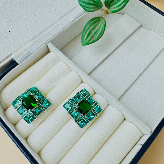 Emerald green square stud earrings with a geometric design, featuring sparkling gemstones set in a silver-tone metal frame, displayed in a cream-colored jewelry box.