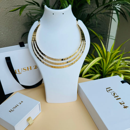 A close-up shot of a large, vintage-inspired gold necklace. The necklace features numerous elongated, slightly curved metallic elements radiating outwards from a central band. It is displayed on a smooth white jewelry bust. In the background, partially visible, are white shopping bags with black handles and the text "LUSH 24" in gold lettering, and a small white jewelry box with the same branding. A blurred view of green tropical foliage is also present.