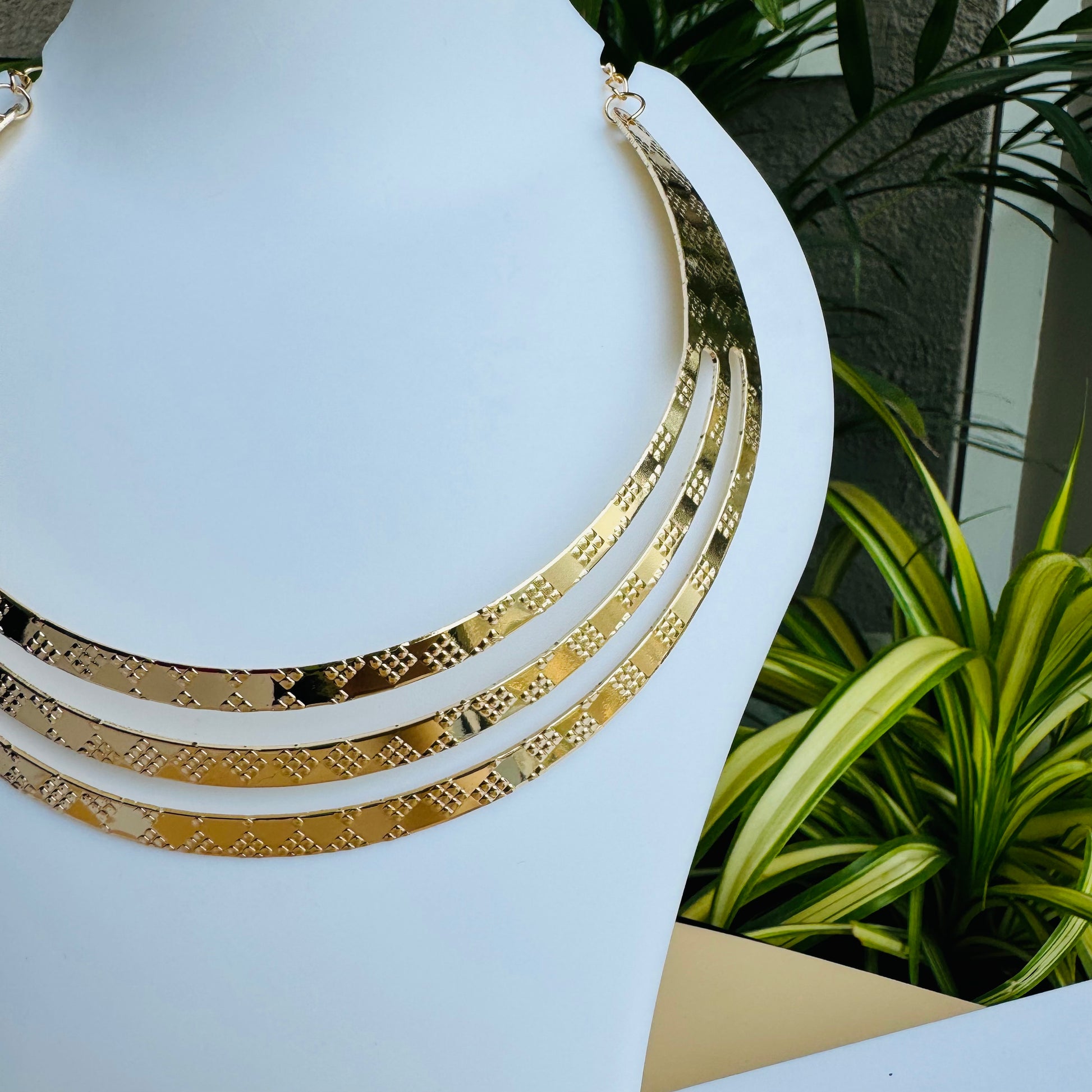 A close-up shot of a large, vintage-inspired gold necklace. The necklace features numerous elongated, slightly curved metallic elements radiating outwards from a central band. It is displayed on a smooth white jewelry bust. In the background, partially visible, are white shopping bags with black handles and the text "LUSH 24" in gold lettering, and a small white jewelry box with the same branding. A blurred view of green tropical foliage is also present.