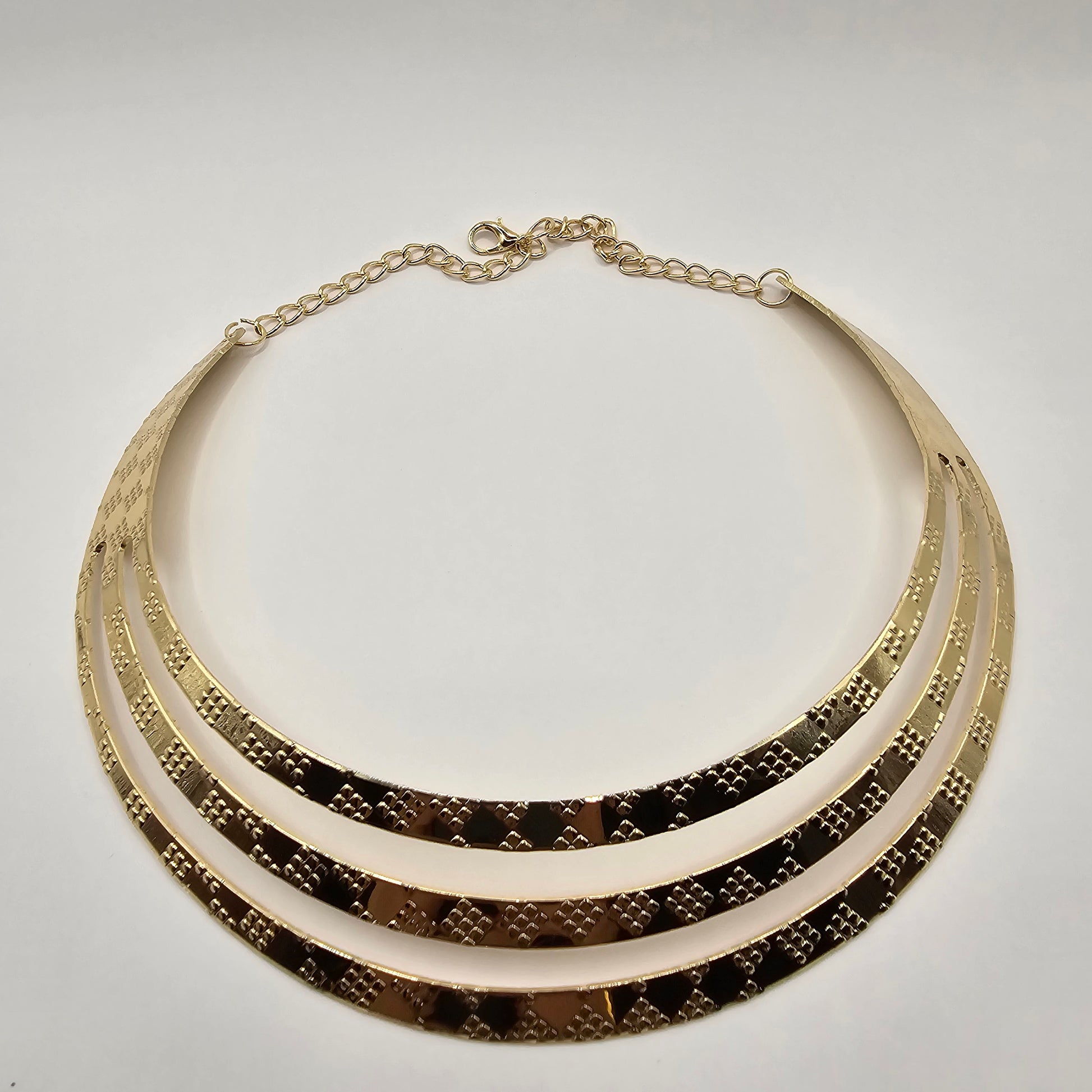 A close-up shot of a large, vintage-inspired gold necklace. The necklace features numerous elongated, slightly curved metallic elements radiating outwards from a central band. It is displayed on a smooth white jewelry bust. In the background, partially visible, are white shopping bags with black handles and the text "LUSH 24" in gold lettering, and a small white jewelry box with the same branding. A blurred view of green tropical foliage is also present.