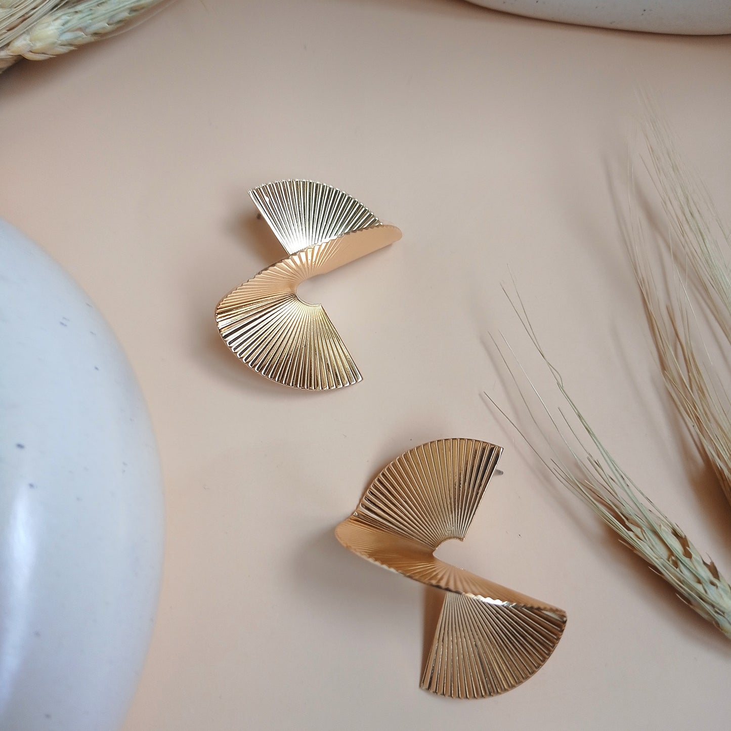 Modern gold statement earrings with a unique spiral fan design, featuring fine ridged detailing, displayed on a neutral beige background with natural accents