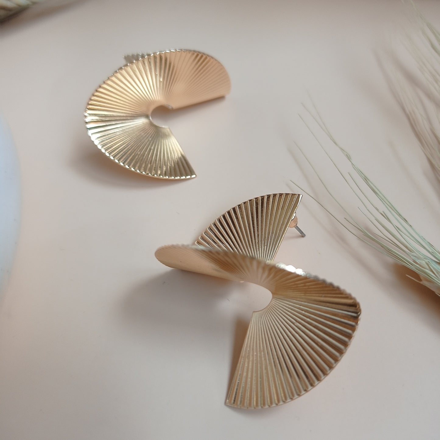 Modern gold statement earrings with a unique spiral fan design, featuring fine ridged detailing, displayed on a neutral beige background with natural accents