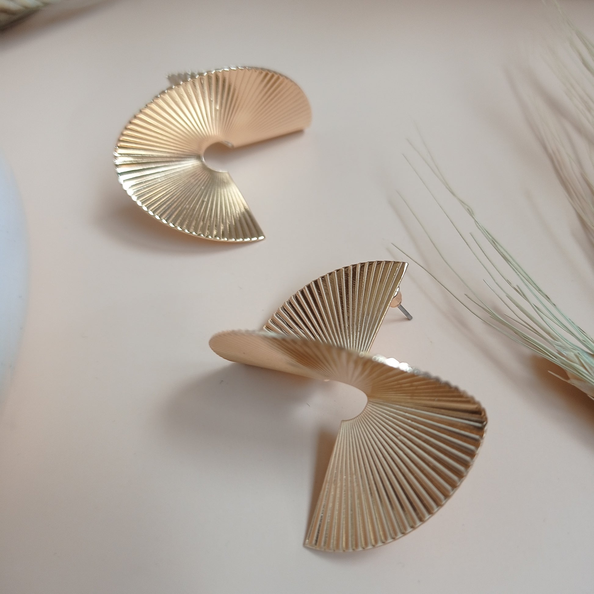 Modern gold statement earrings with a unique spiral fan design, featuring fine ridged detailing, displayed on a neutral beige background with natural accents