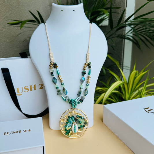 A statement necklace displayed on a white bust stand, featuring dual strands of turquoise and gold-toned beads, interspersed with faceted stones. The necklace has an intricate circular pendant with gold filigree, green gemstones, and a central shell-like embellishment. Luxury packaging with "LUSH 24" branding surrounds the display, set against a backdrop of green plants