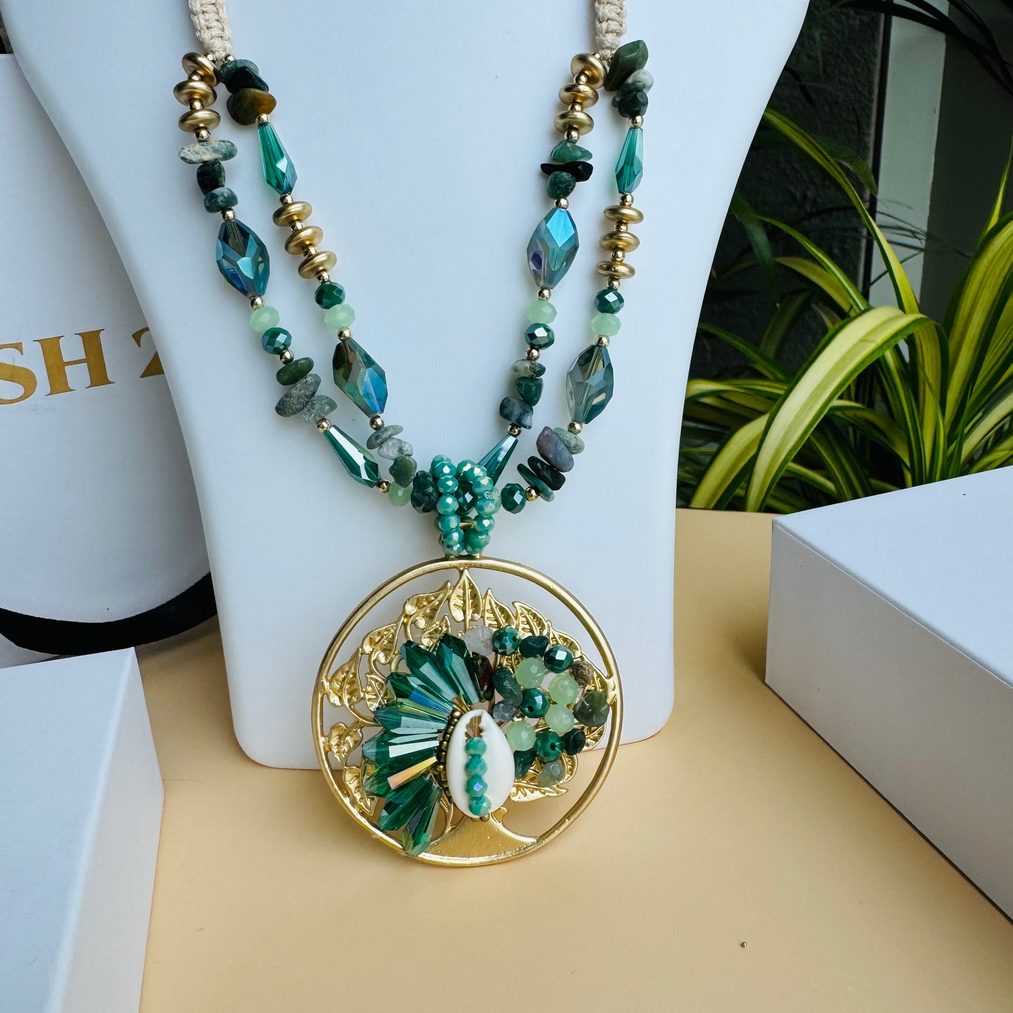 A statement necklace displayed on a white bust stand, featuring dual strands of turquoise and gold-toned beads, interspersed with faceted stones. The necklace has an intricate circular pendant with gold filigree, green gemstones, and a central shell-like embellishment. Luxury packaging with "LUSH 24" branding surrounds the display, set against a backdrop of green plants